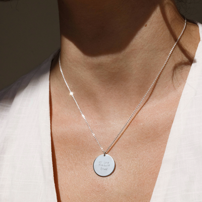 A close up lifestyle shot of a person's neck wearing a delicate silver pendant chain with a large round disc, featuring engraved text on the front.