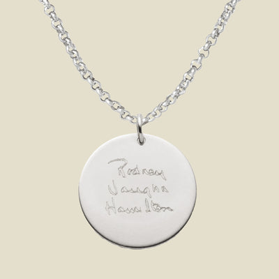 A silver chain necklace with a round silver pendant engraved with handwritten script, shown against a solid light beige background.