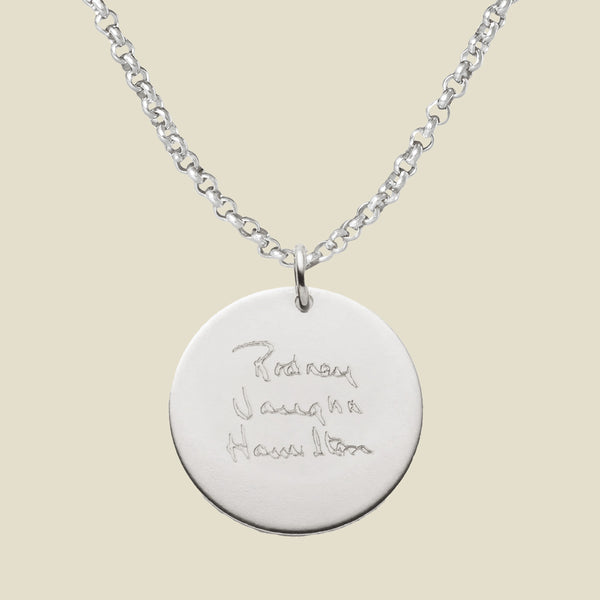 A silver chain necklace with a round silver pendant engraved with handwritten script, shown against a solid light beige background.