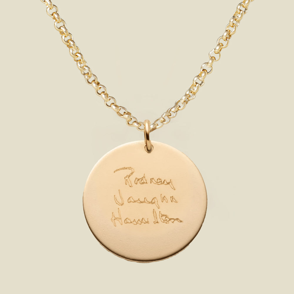 A gold chain necklace with a round gold pendant engraved with handwritten script, shown against a solid light beige background.