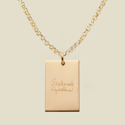 Gold necklace with a rectangular engraved pendant featuring handwritten text on a beige background