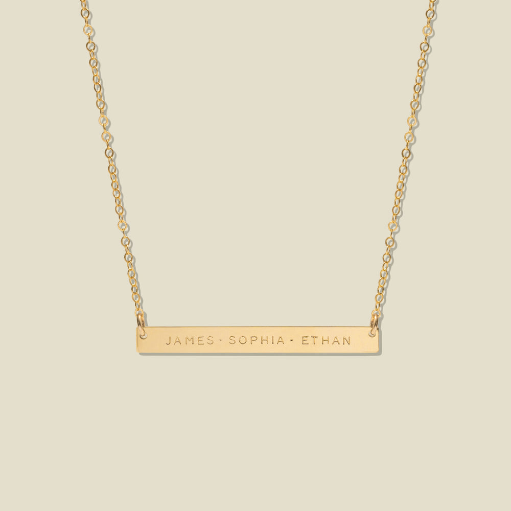 A gold bar necklace with three engraved names, 'JAMES', 'SOPHIA', and 'ETHAN', suspended from a delicate gold chain against a solid light beige background.