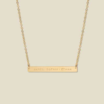 A gold bar necklace with three engraved names, 'JAMES', 'SOPHIA', and 'ETHAN', suspended from a delicate gold chain against a solid light beige background.