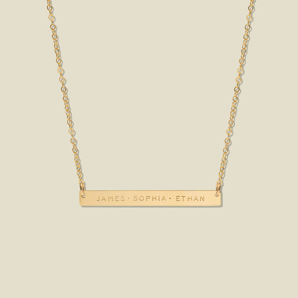 A gold bar necklace with three engraved names, 'JAMES', 'SOPHIA', and 'ETHAN', suspended from a delicate gold chain against a solid light beige background.