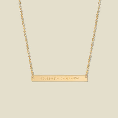 A gold bar necklace with engraved coordinates, 40.6892'N and 74.0445'W, suspended from a delicate gold chain against a solid light beige background.