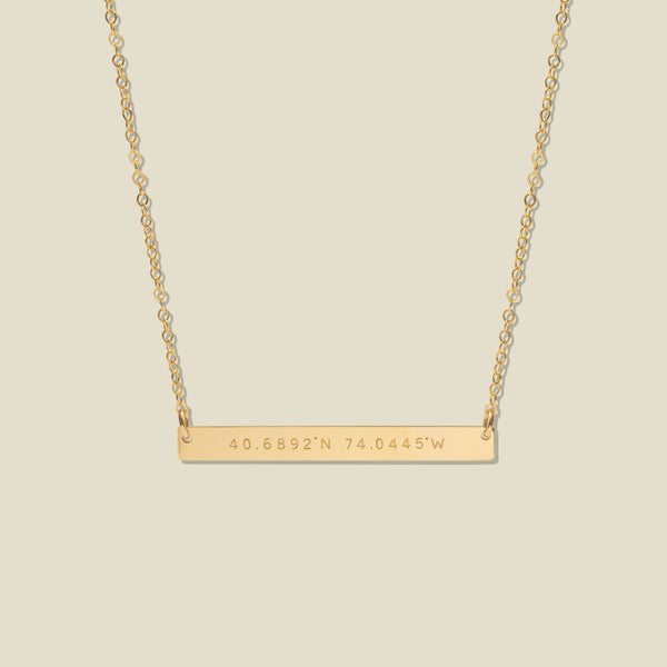 A gold bar necklace with engraved coordinates, 40.6892'N and 74.0445'W, suspended from a delicate gold chain against a solid light beige background.