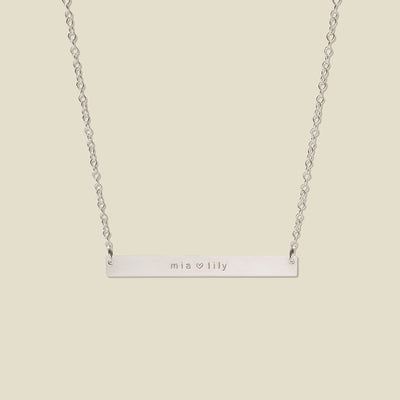 A silver bar necklace with two engraved names, 'mia' and 'lily' separated by a heart, suspended from a delicate silver chain against a solid light beige background.