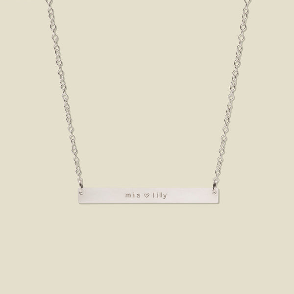 A silver bar necklace with two engraved names, 'mia' and 'lily' separated by a heart, suspended from a delicate silver chain against a solid light beige background.