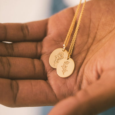 The palm of a hand holding two delicate gold pendant chain necklaces, showcasing the flat oval disc charms engraved with a stem of flowers.