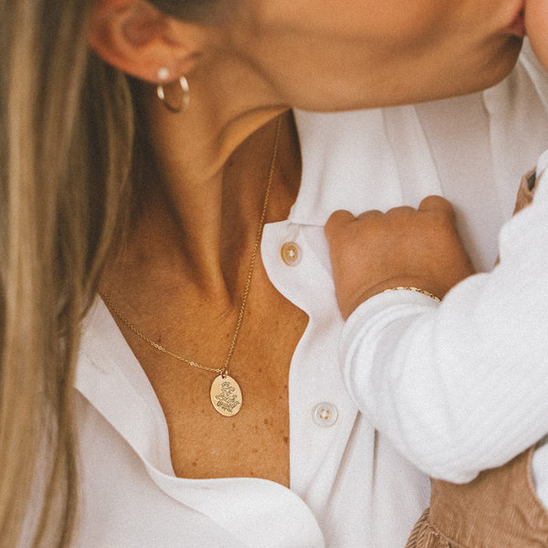 A close-up of a woman's chest holding a small child, showcasing a delicate gold pendant chain necklace with a floral engraved flat oval disc.