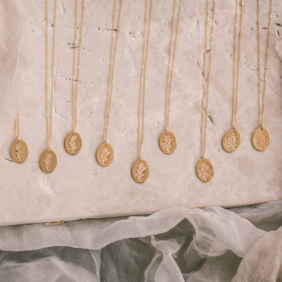 Nine delicate gold pendant necklaces, each with a flat oval disc featuring a floral motif, laying on a beige stone surface. From left to right, the amount of flowers on each engraved disc goes up from one flower to nine.