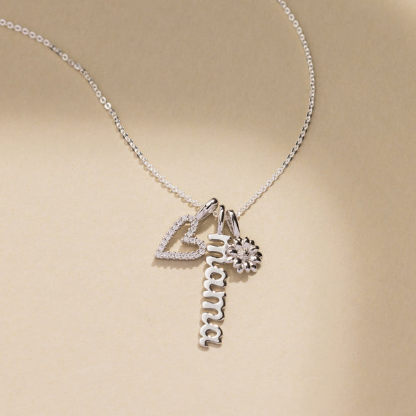 A silver pendant necklace with a heart charm covered in clear stones, a vertical nameplate 'mama' charm in script font, and a small daisy charm.