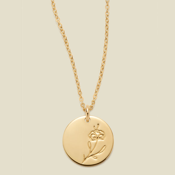 A gold chain necklace with a round gold disc pendant featuring an engraved violet flower design, displayed against a solid light beige background.