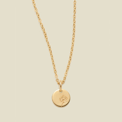 A gold chain necklace with a round gold disc pendant featuring an engraved violet flower design, displayed against a solid light beige background.