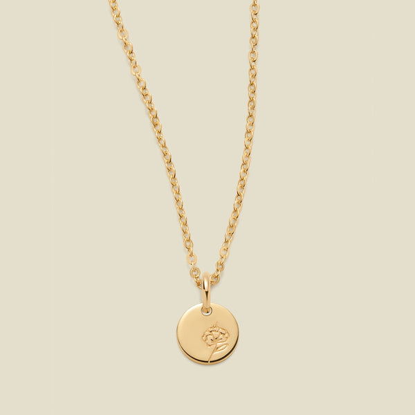 A gold chain necklace with a round gold disc pendant featuring an engraved violet flower design, displayed against a solid light beige background.