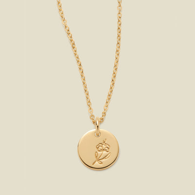 A gold chain necklace with a round gold disc pendant featuring an engraved violet flower design, displayed against a solid light beige background.