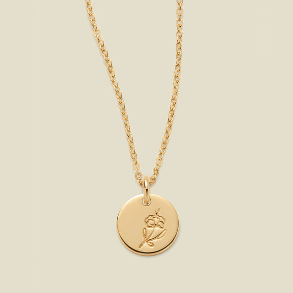 A gold chain necklace with a round gold disc pendant featuring an engraved violet flower design, displayed against a solid light beige background.