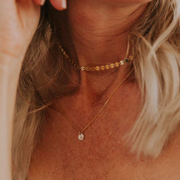 A close-up of a woman wearing a gold choker made up of round flat discs layered with a delicate gold pendant chain featuring a small round flat disc charm. The disc is engraved with a floral design.