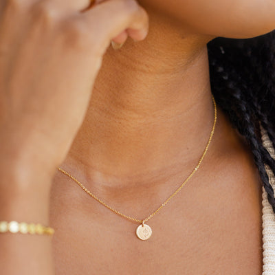A close-up of a woman wearing a delicate gold pendant chain necklace with a round flat disc charm. The disc is engraved with a violet flower design.