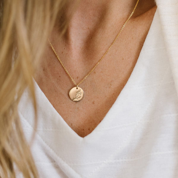 A close-up of a woman in a white v-neck shirt wearing a delicate gold pendant chain with a round flat disc charm. The charm is engraved with a violet flower design.