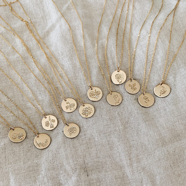 Twelve gold pendant chain necklaces, each with a round flat disc engraved with various floral motifs, laying on a light beige linen background.