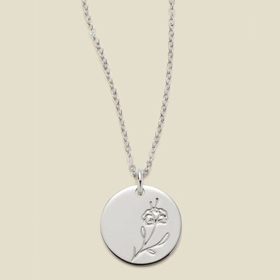 A silver chain necklace with a round silver disc pendant featuring an engraved violet flower design, displayed against a solid light beige background.