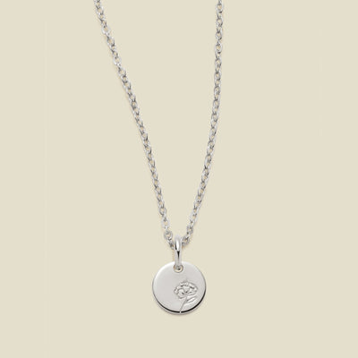 A silver chain necklace with a round silver disc pendant featuring an engraved violet flower design, displayed against a solid light beige background.