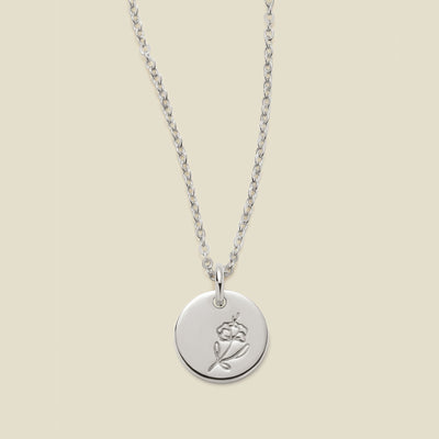 A silver chain necklace with a round silver disc pendant featuring an engraved violet flower design, displayed against a solid light beige background.