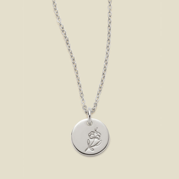 A silver chain necklace with a round silver disc pendant featuring an engraved violet flower design, displayed against a solid light beige background.