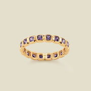 A gold eternity ring featuring a continuous row of square cut purple gemstones, shown on a solid light beige background.
