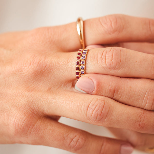 A person's hand wearing two stacked gold eternity rings, each with either purple or dark red square cut gemstones in a single continuous row.