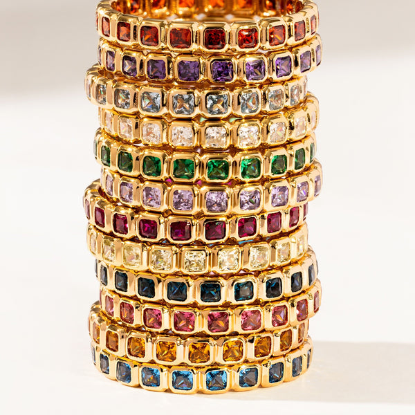 Twelve stacked gold eternity rings, each with a different color of square cut gemstones in a single continuous row, shown on a bright white background.