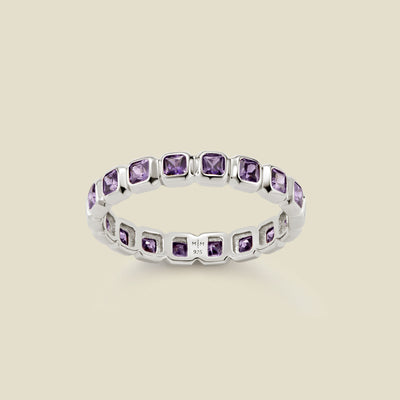A silver eternity ring featuring a continuous row of square cut purple gemstones, shown on a solid light beige background.
