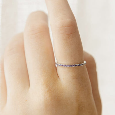 A close-up of a hand with a thin silver ring, featuring a single row of small round-cut purple gemstones across the front, set against a bright white background.