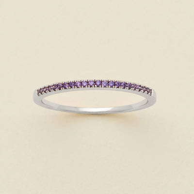 A silver ring featuring a single row of small round-cut dark purple gemstones, photographed on a clean, solid, warm off-white background.