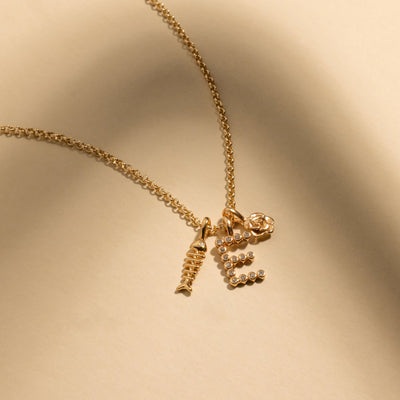 A gold chain necklace with a gold fish skeleton charm, an intial 'E' charm made up of small round clear gemstones, and a gold rose charm.