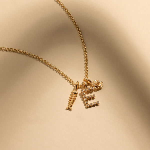 A gold chain necklace with a gold fish skeleton charm, an intial 'E' charm made up of small round clear gemstones, and a gold rose charm.