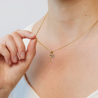 A person wearing a gold chain necklace with a gold fish skeleton charm and a bezel-set clear teardrop gemstone charm.