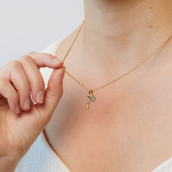 A person wearing a gold chain necklace with a gold fish skeleton charm and a bezel-set clear teardrop gemstone charm.