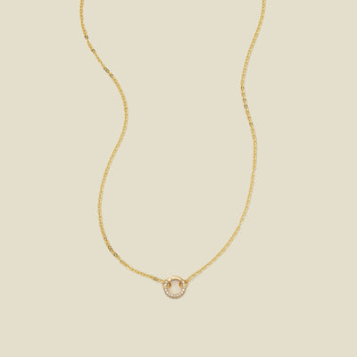 A delicate gold chain necklace with a gold round ring covered in clear gemstones in the center, set against a solid beige background.