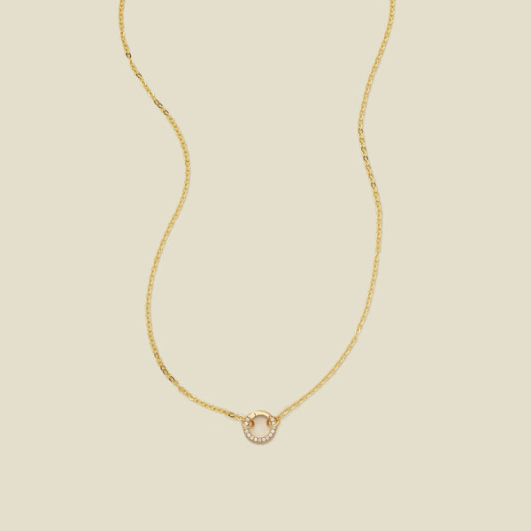A delicate gold chain necklace with a gold round ring covered in clear gemstones in the center, set against a solid beige background.