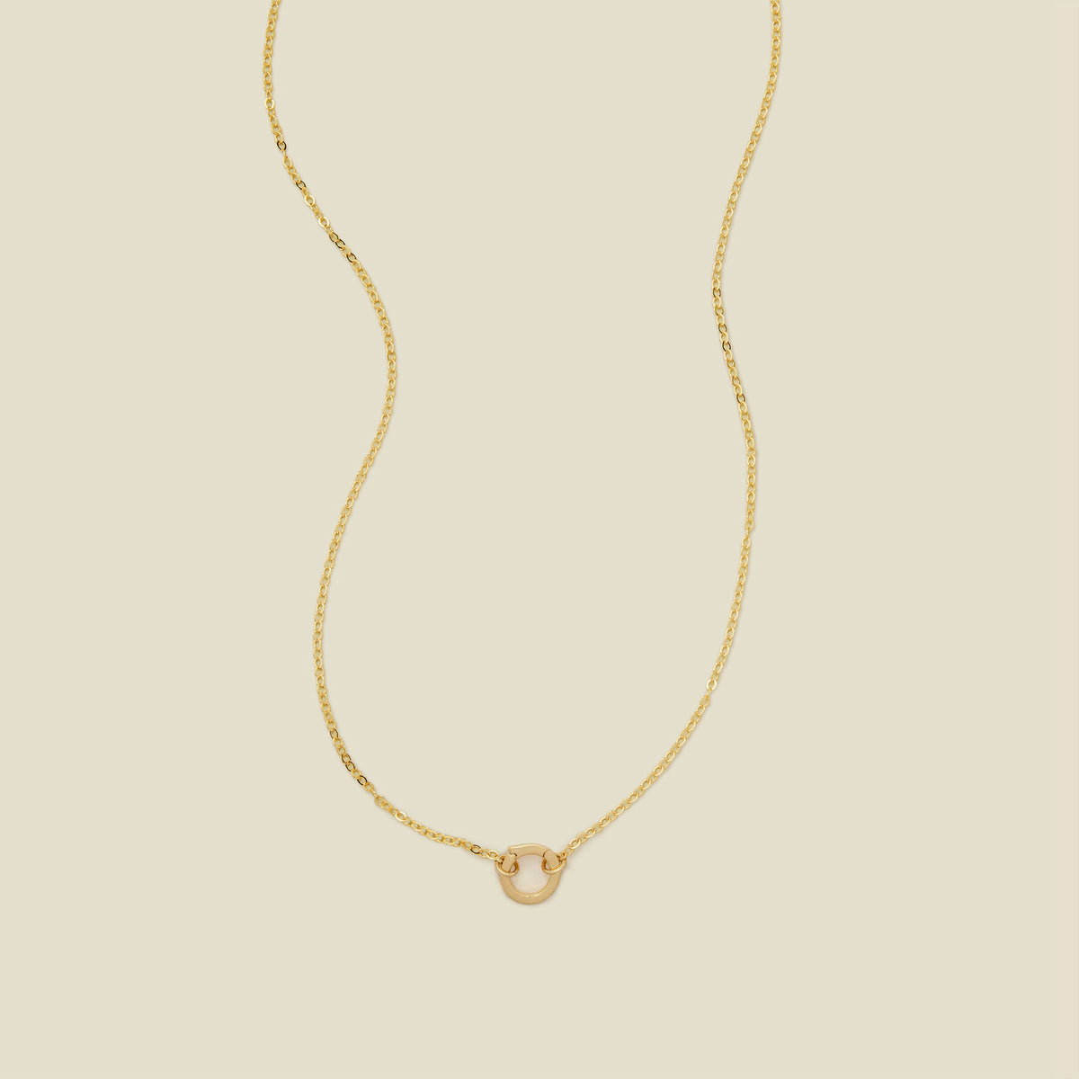 A delicate gold chain necklace with a polished gold round ring in the center, set against a solid beige background.