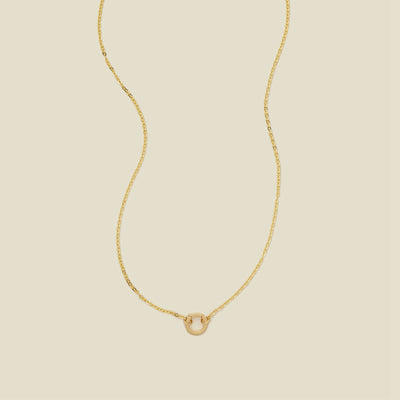 A delicate gold chain necklace with a polished gold round ring in the center, set against a solid beige background.