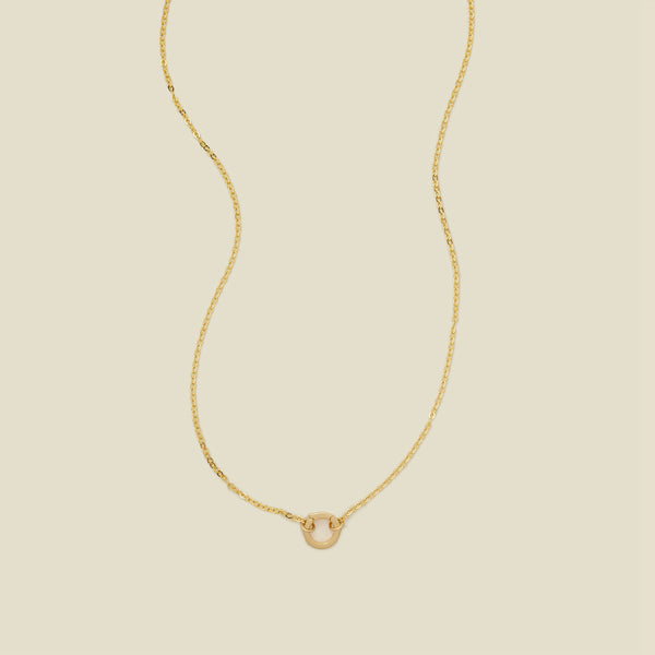 A delicate gold chain necklace with a polished gold round ring in the center, set against a solid beige background.