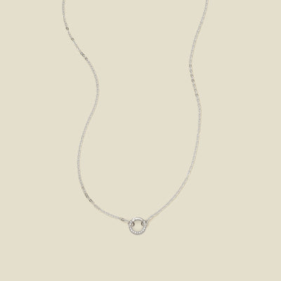 A delicate silver chain necklace with a silver ring covered in clear accents in the center, set against a solid beige background.