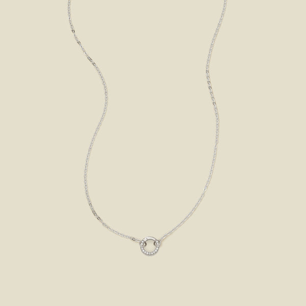 A delicate silver chain necklace with a silver ring covered in clear accents in the center, set against a solid beige background.