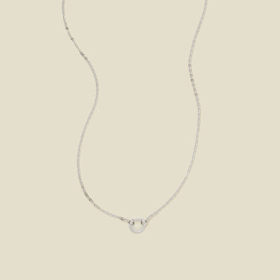 A delicate silver chain necklace with a polished silver ring in the center, set against a solid beige background.