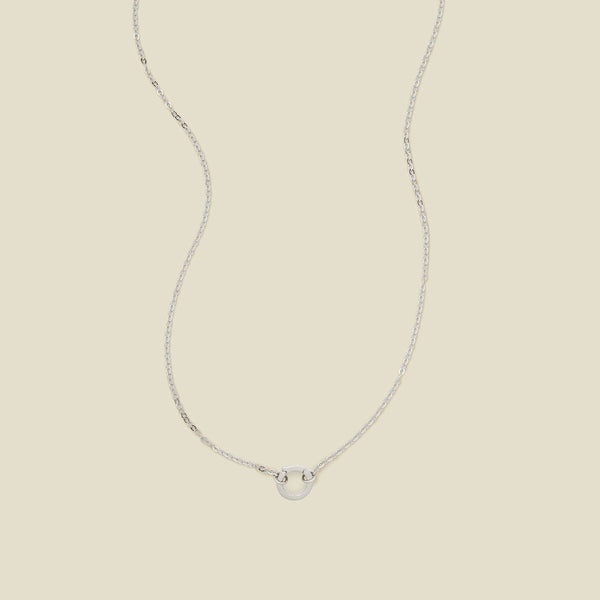 A delicate silver chain necklace with a polished silver ring in the center, set against a solid beige background.