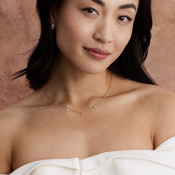 A close-up of a person's chest wearing a dainty gold chain necklace with three dangling clear gemstone flowers and two dangling round clear stones, paired with a white off the shoulder top and set against a light brown textured backdrop.
