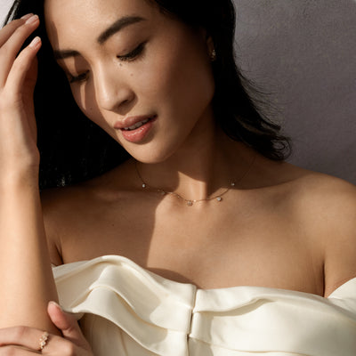 A close-up of a woman wearing a white ruffled off the shoulder top and delicate gold chain necklace with three dangling flowers and two dangling round clear stones, set against a dark grey background.